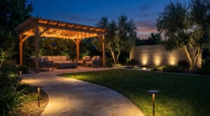 Modern outdoor patio at dusk with a string-lit pergola, comfortable seating, stone pathway with path lights, and uplights on trees.