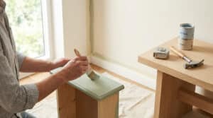 Mains d'une personne peignant un petit meuble en bois vert d'eau. Sur une table, marteau, mètre et pots de peinture.